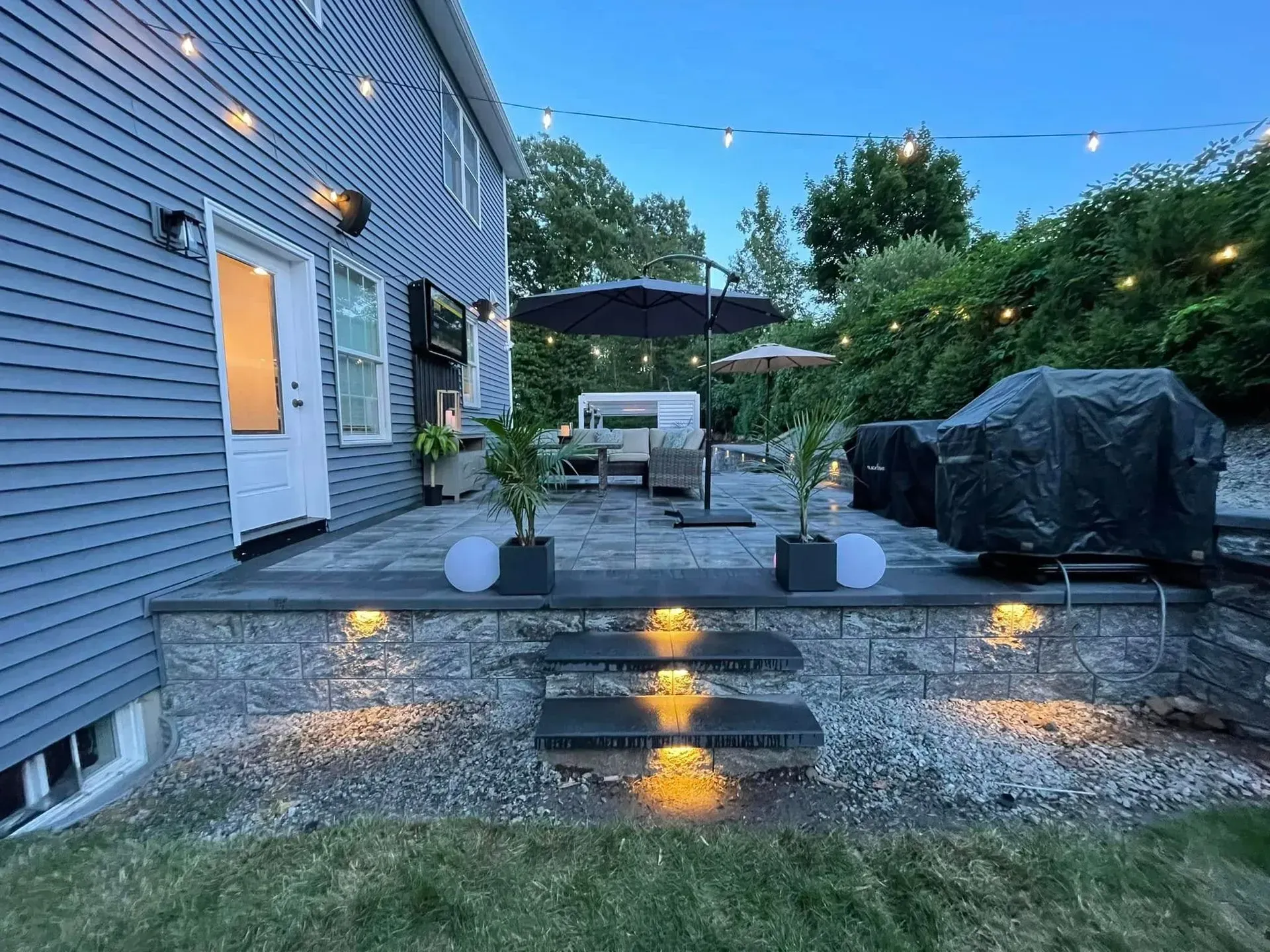 Backyard patio at dusk with string lights, grill, umbrella, and a lounge area.