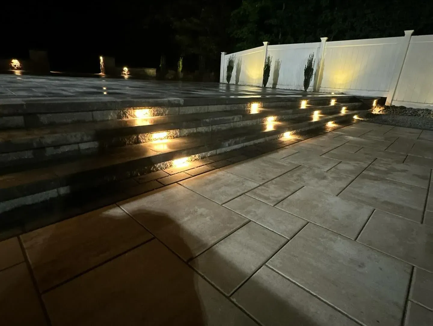 Stone patio with illuminated steps and a white fence at night.