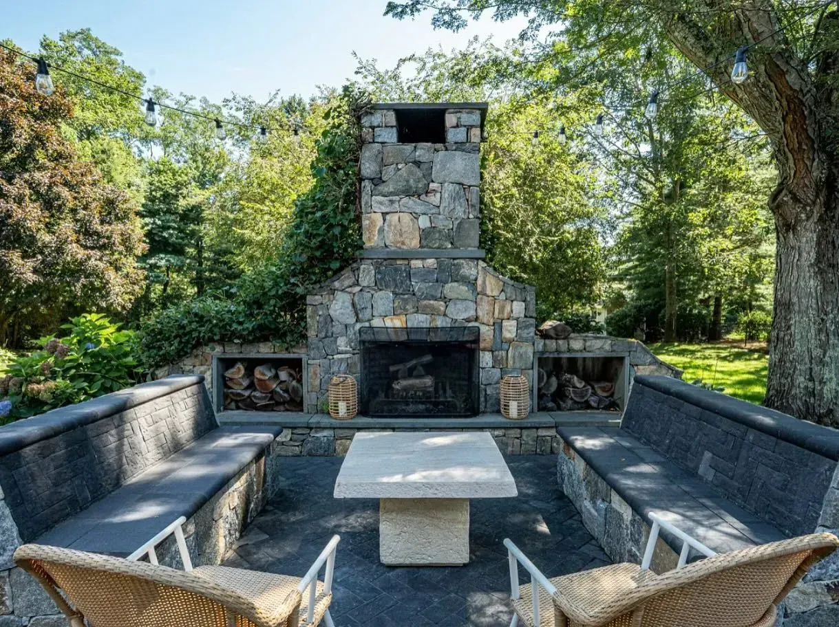 Outdoor stone fireplace with built-in seating and table; surrounded by trees and greenery.