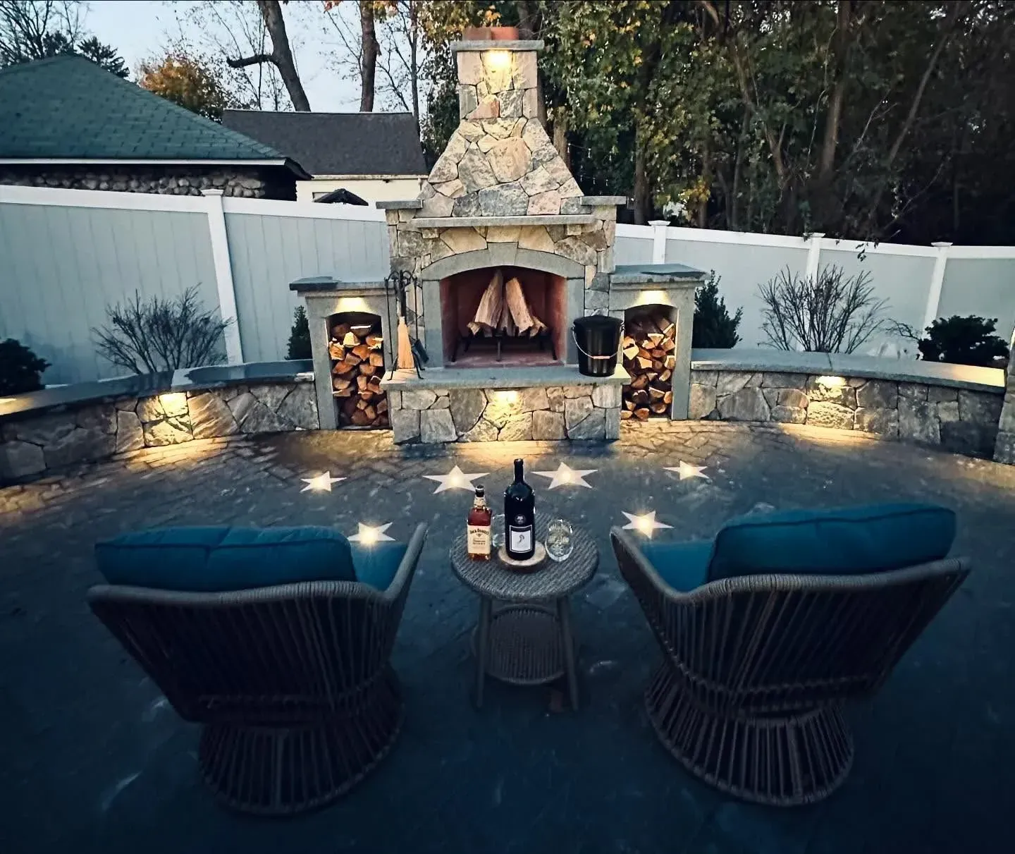 Outdoor fireplace with chairs, side table with wine, and glowing lights.