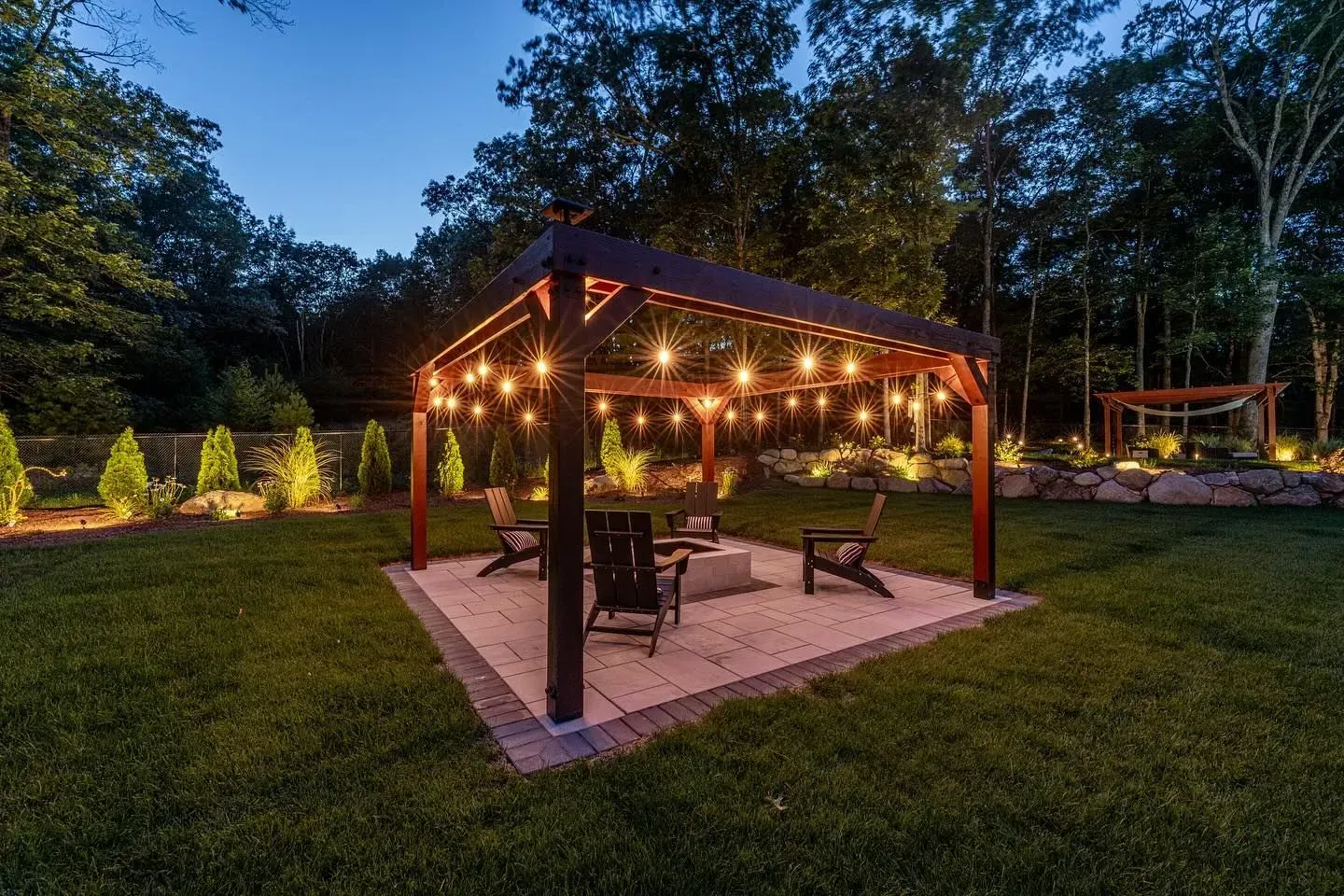 A backyard pergola with string lights, chairs, and a fire pit, surrounded by trees at dusk.