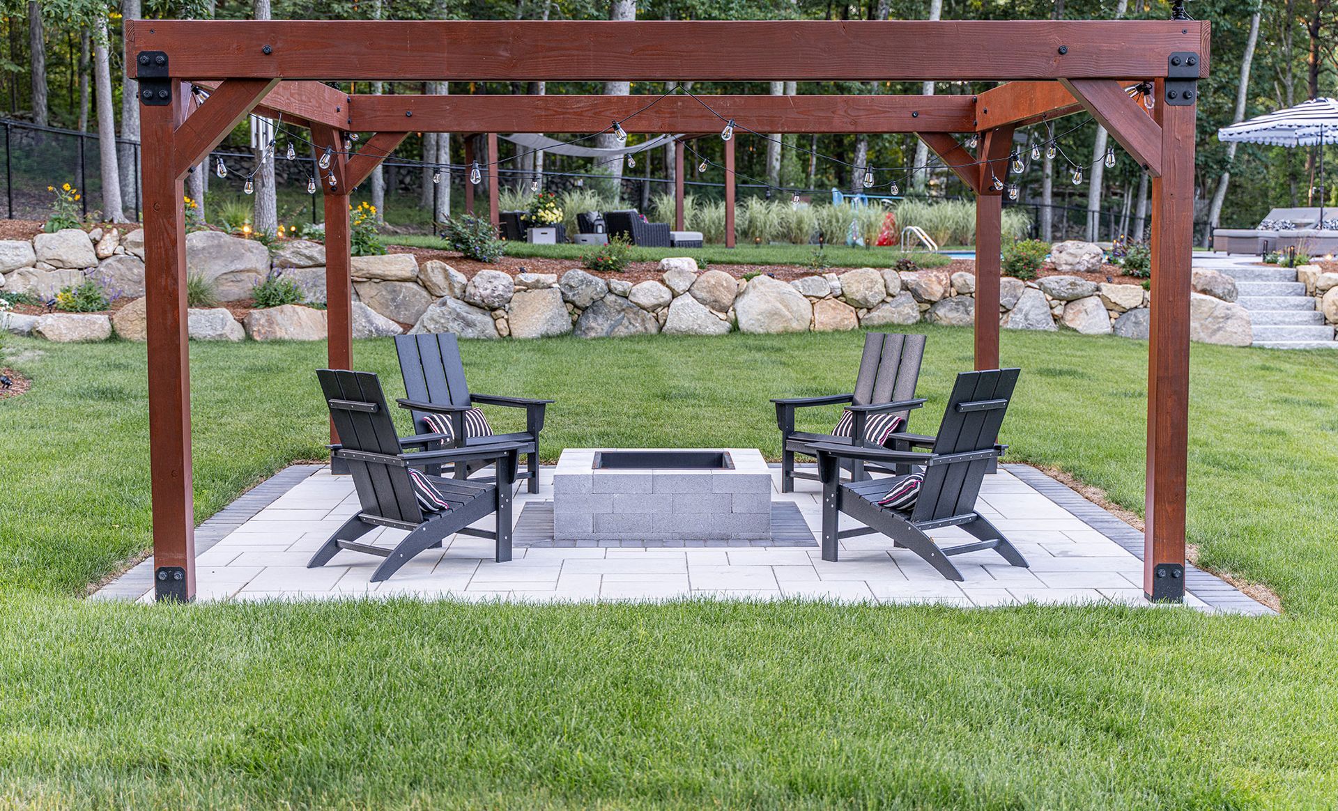 Pergola over a fire pit with four black chairs on a paver patio in a backyard setting.