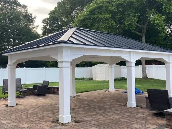 White gazebo with black metal roof on a brick patio in a backyard setting.