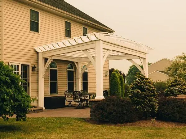 White pergola attached to a beige house, over a patio with black furniture, green bushes in the yard.