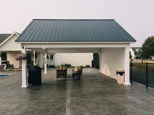 Covered outdoor patio with dark gray metal roof, seating, and a grill. White support columns and siding.