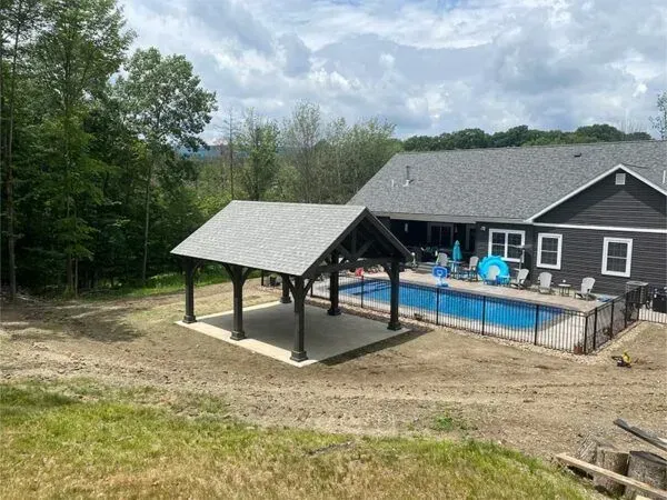 Pavilion with gray roof over a concrete patio next to a pool and dark house on a sunny day.