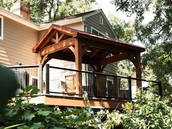 Wooden deck with covered seating area attached to a house with black railings and surrounding greenery.