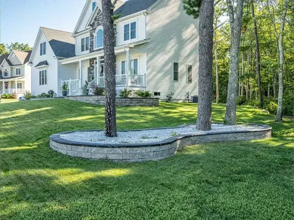 House with a curved stone retaining wall surrounding trees, on a grassy hill.