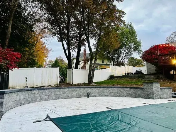 Curved stone wall with a pool cover in the foreground and white fence, trees and a house in the background.