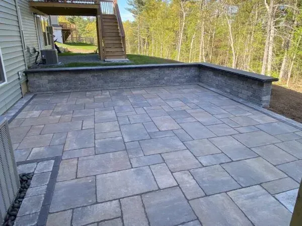Stone patio with retaining wall next to a house and stairs leading up to a deck, with trees in the background.