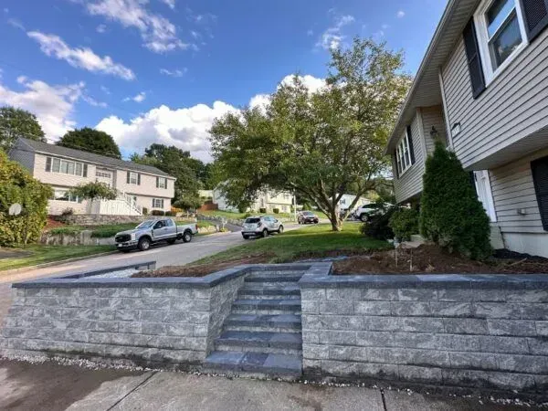 Gray brick retaining wall with steps leading up to a house on a sunny day.