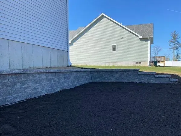 Grey retaining wall with steps, black soil in front, light green house background.