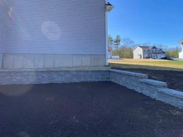 Exterior view of a house with a concrete foundation, retaining wall, and freshly laid dark soil. Sunny day.