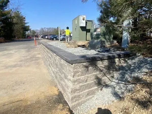 Retaining wall made of gray blocks. Worker near electrical box, asphalt road, and cars.