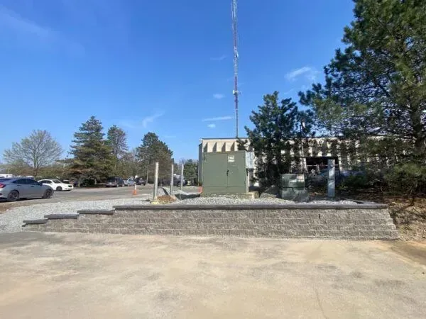 Gray block retaining wall with gravel parking area. Green utility box and radio tower against a blue sky.