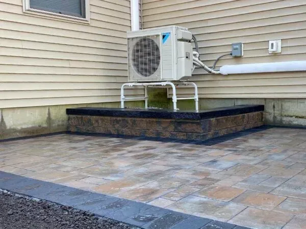 Outdoor air conditioning unit on a stone patio with a concrete border, beside a light-yellow house.
