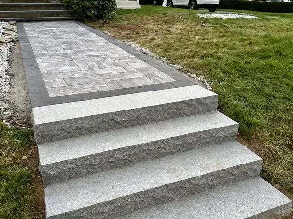Stone steps leading up to a paved walkway with a brick-like pattern, bordered by dark gray stone and lawn.