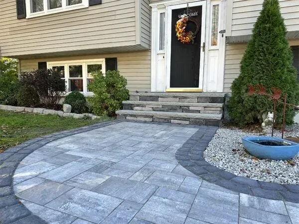 Paver patio and steps leading to a house with a black door, surrounded by landscaping.