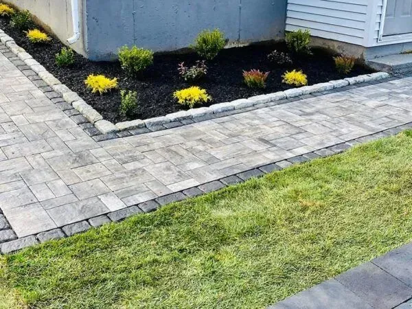 A paved walkway borders a flowerbed with shrubs and yellow flowers, next to a grassy lawn.