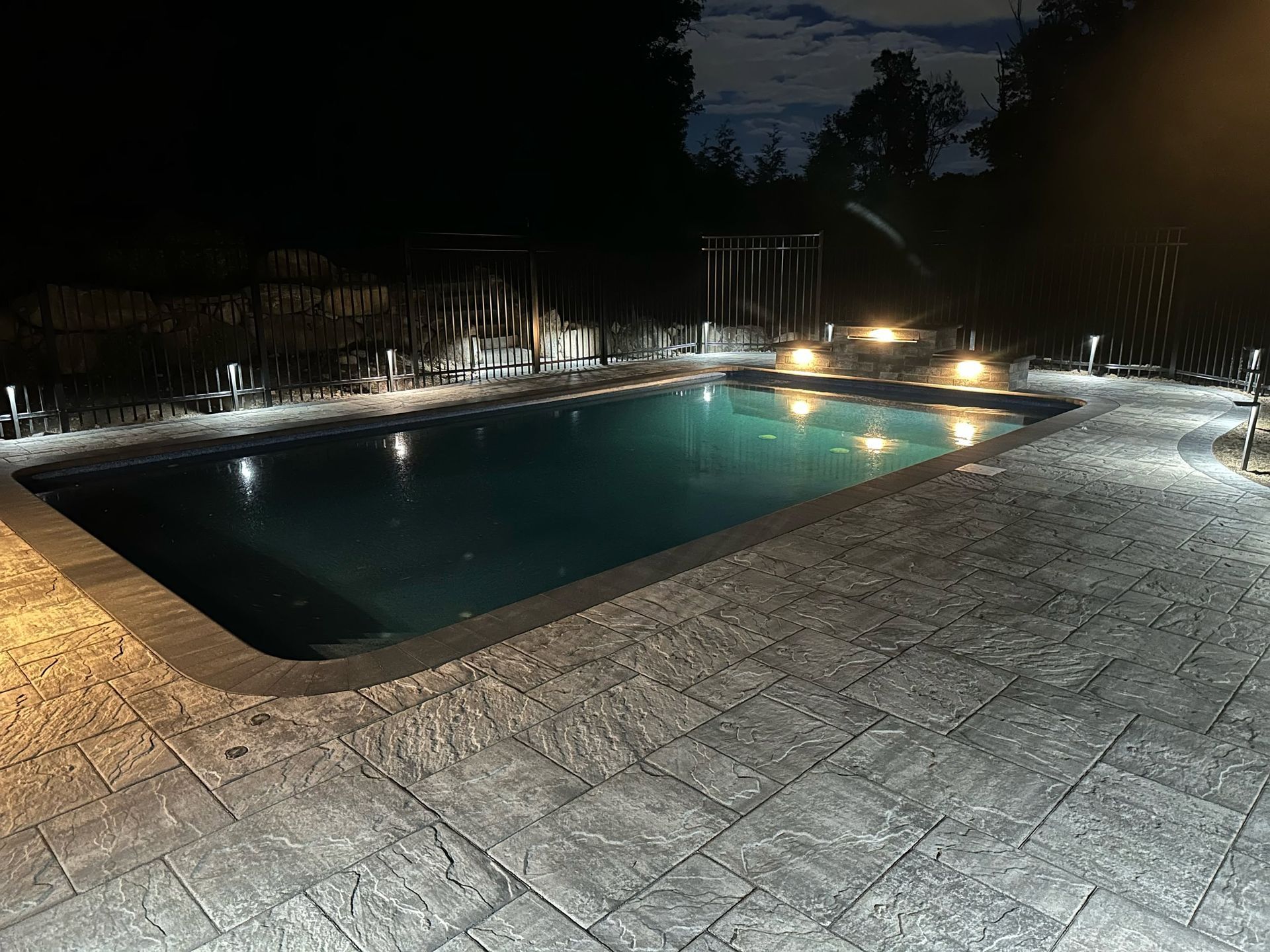 Illuminated backyard swimming pool at night with stone patio and surrounding fence