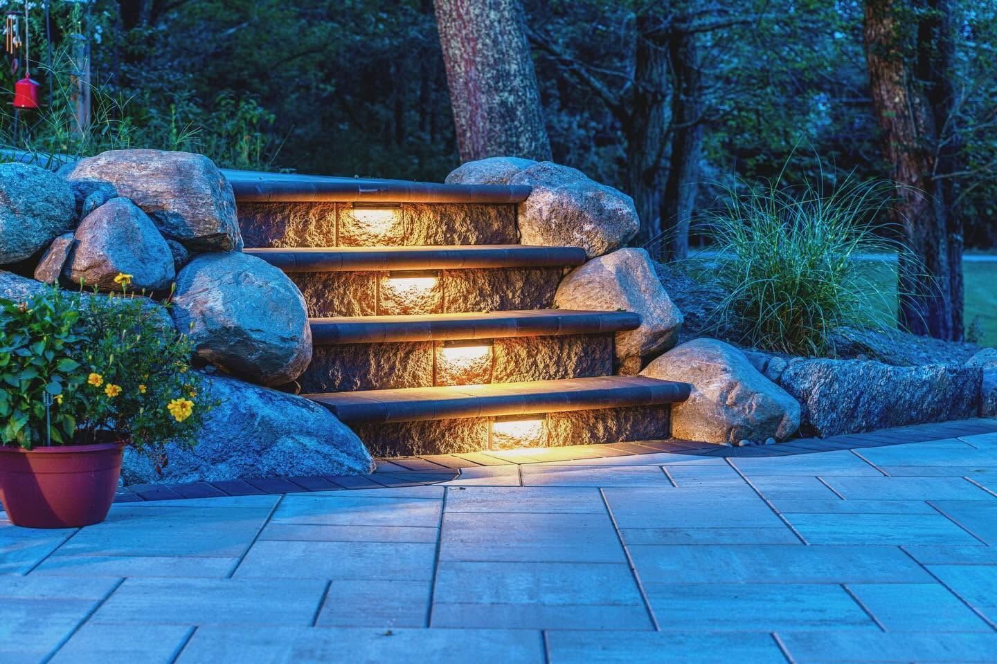 Illuminated stone steps in a landscaped garden at dusk, with potted flowers and surrounding rocks.