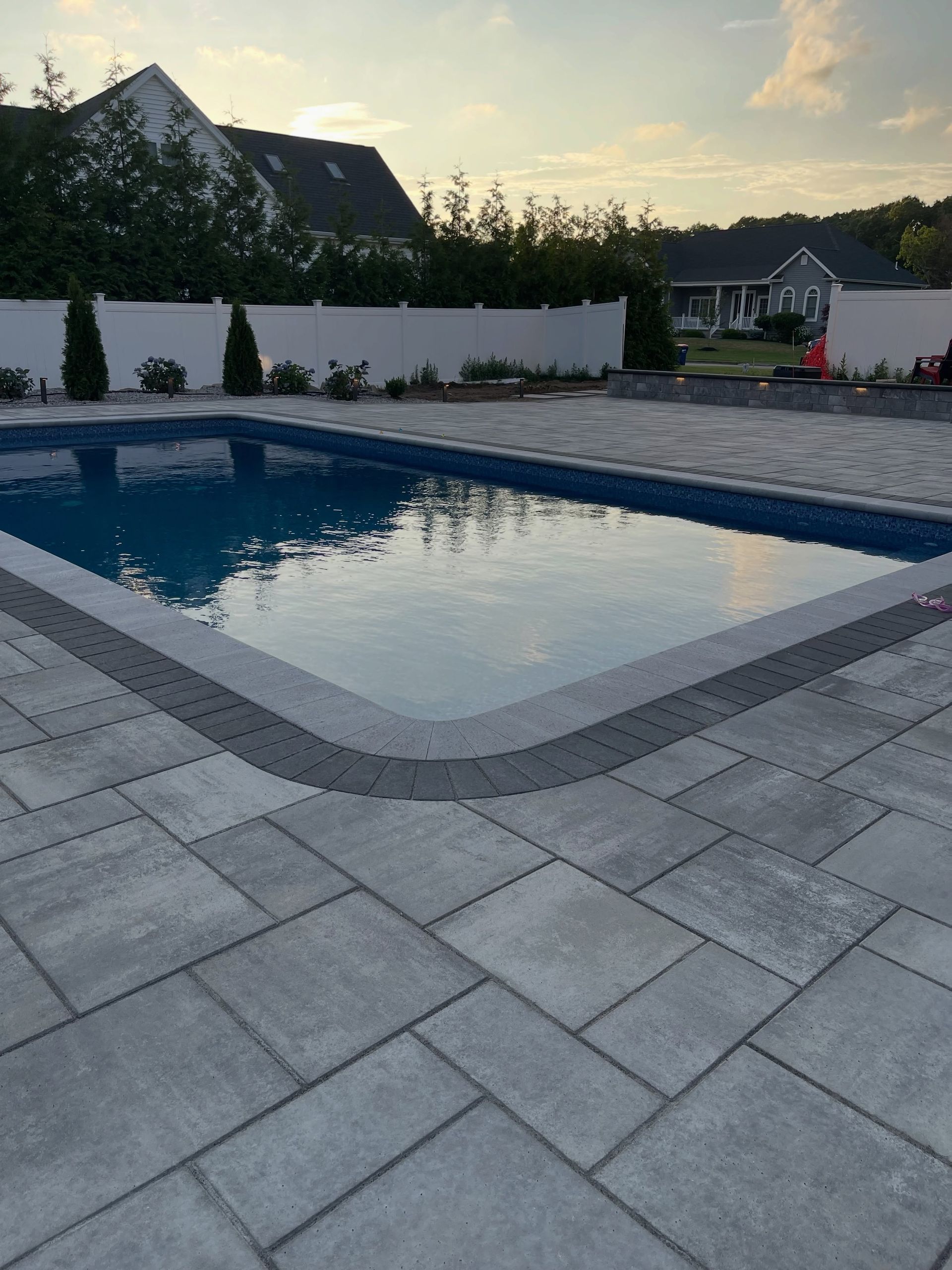 Backyard swimming pool at dusk with white fence, paved patio, and reflections on the water