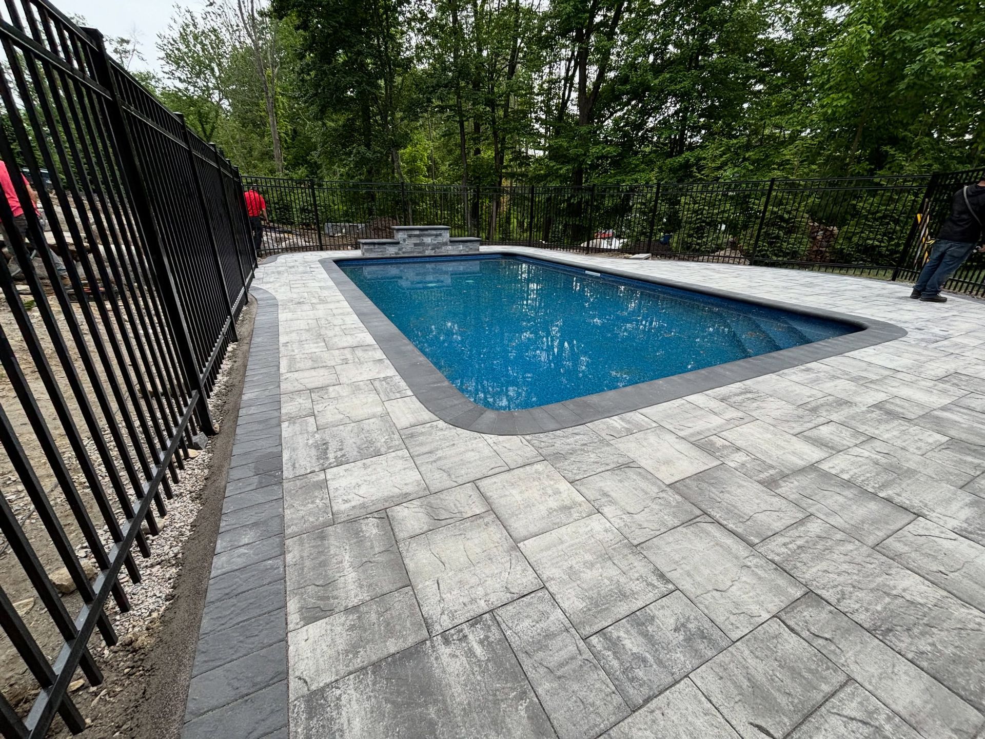 Outdoor swimming pool with blue water, stone deck, and black fence surrounded by trees