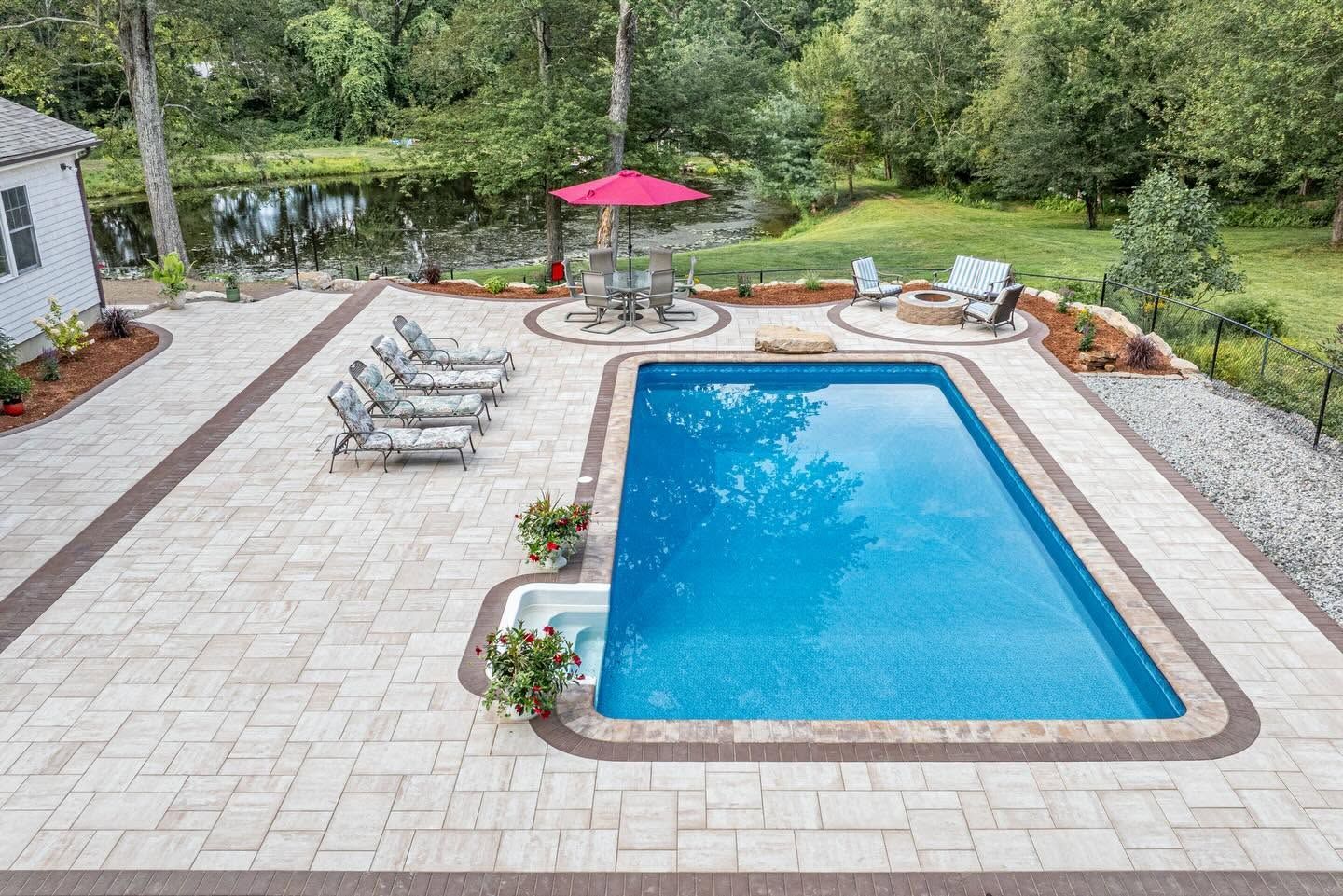 Backyard swimming pool with lounge chairs, stone patio, and red umbrella surrounded by greenery