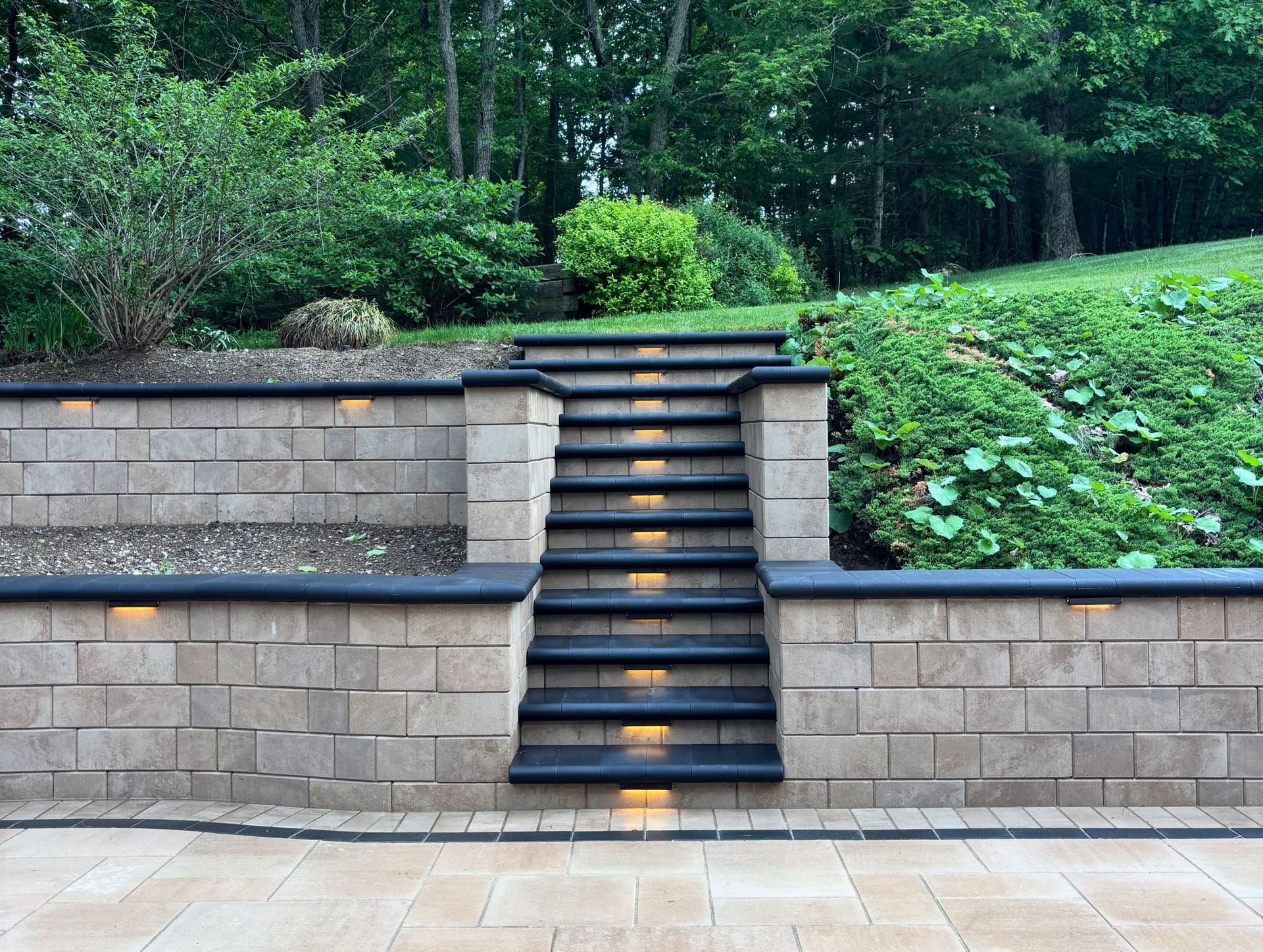 Stone stairs centered in a landscaped retaining wall, leading up to a green wooded area.