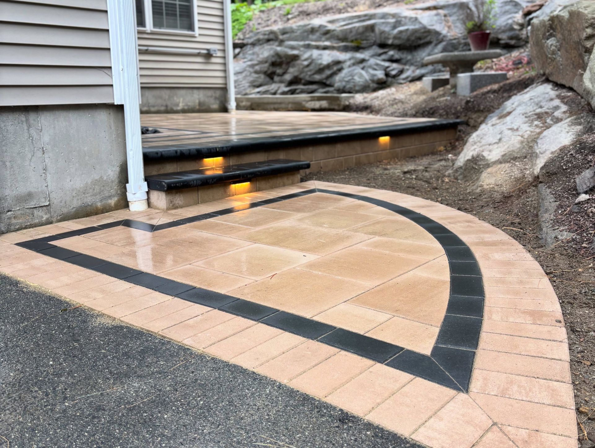 Accessible ramp and walkway with tan paving and black border beside a house and rock steps