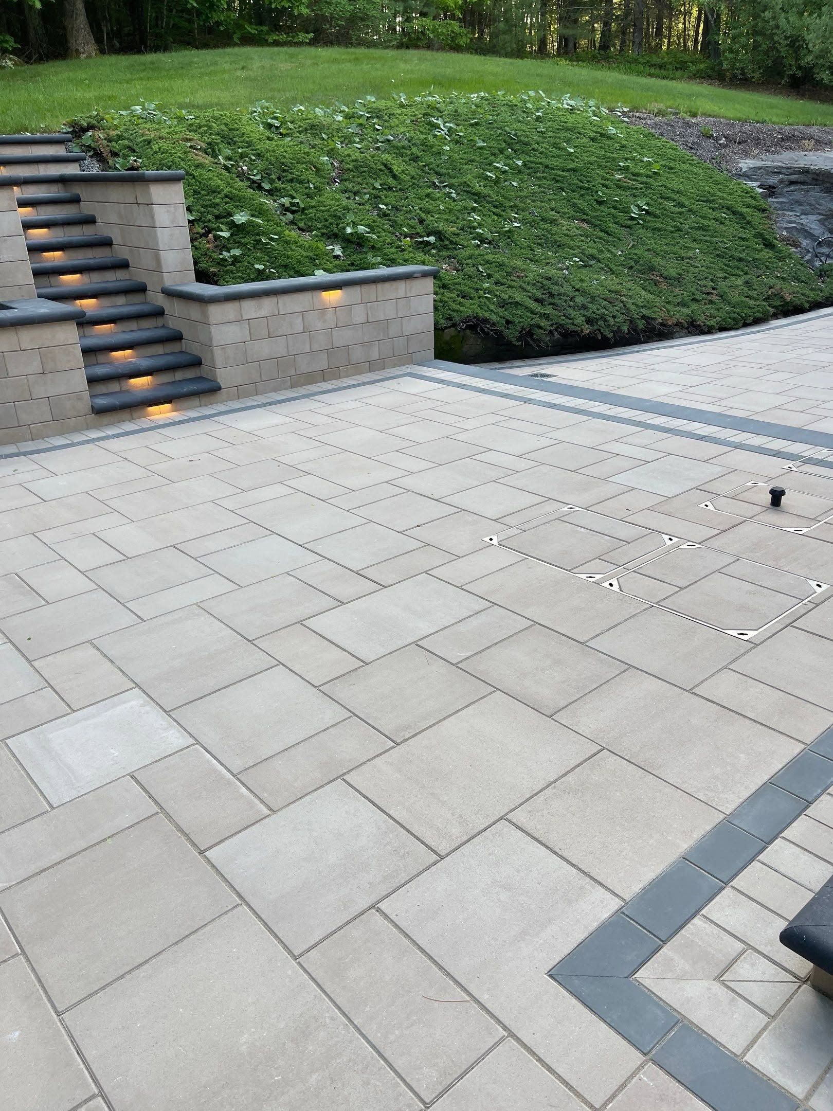 Stone patio with stairs, low brick walls, and dense green hedges by a paved walkway