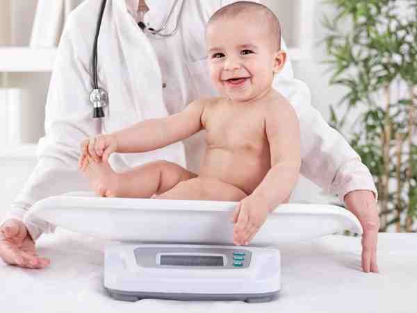 Smiling baby sitting on a white scale in a doctor’s office