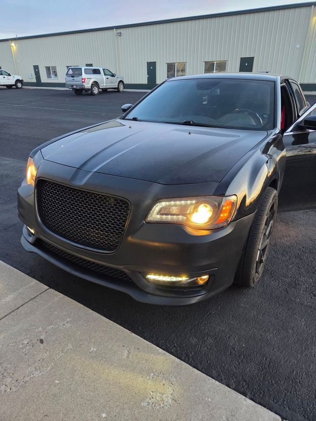 Black Chrysler sedan parked in a lot at dusk, front left headlight on.
