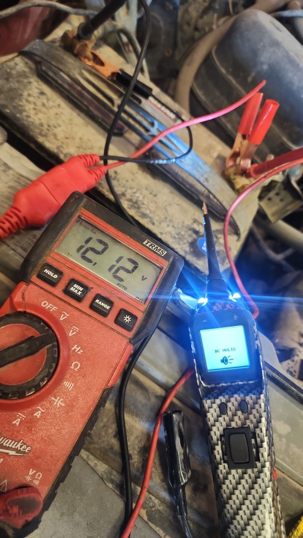 Digital multimeter testing wires beside a small blue-lit circuit board on a cluttered workbench.