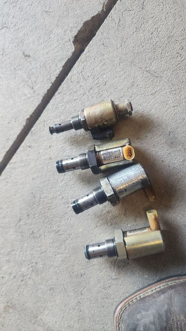 Four small metal automotive solenoids/valves lying on a concrete floor near a shoe tip