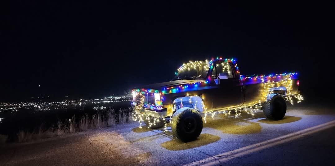 Jeep covered in colorful holiday lights driving at night on a road with city lights in the distance