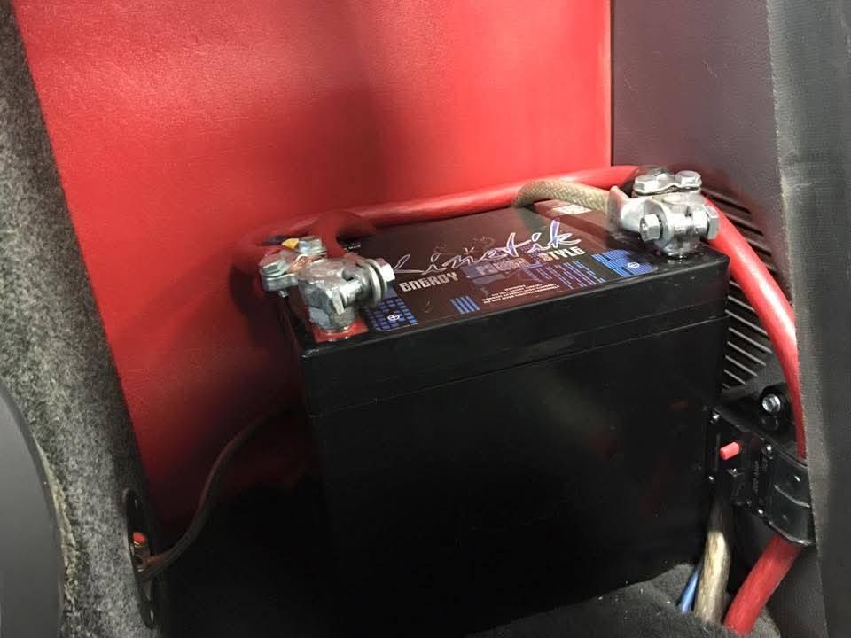 Black battery with red cables mounted in a vehicle compartment against a red panel