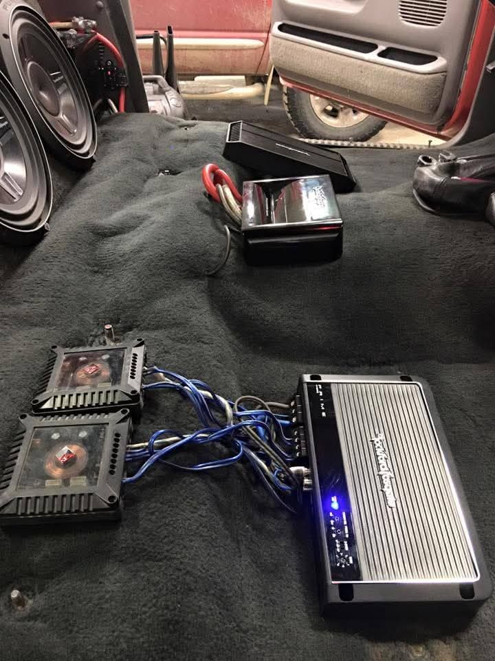 Car audio setup with two amps, blue wiring, and a small speaker on a carpeted car floor