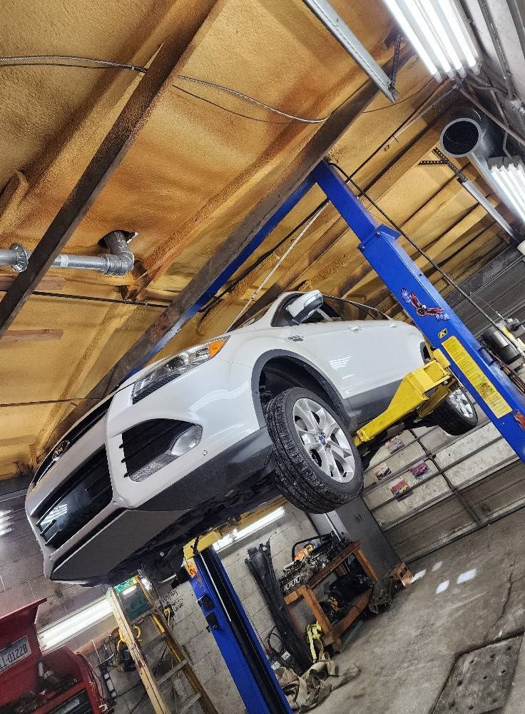 White car lifted on a blue hydraulic lift inside an auto repair shop with a wooden ceiling.
