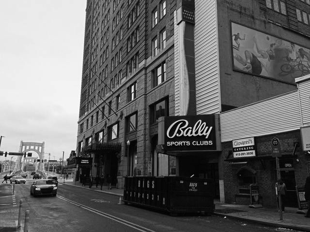 Bally Sports Club building on city street with parked cars and bridge in background.