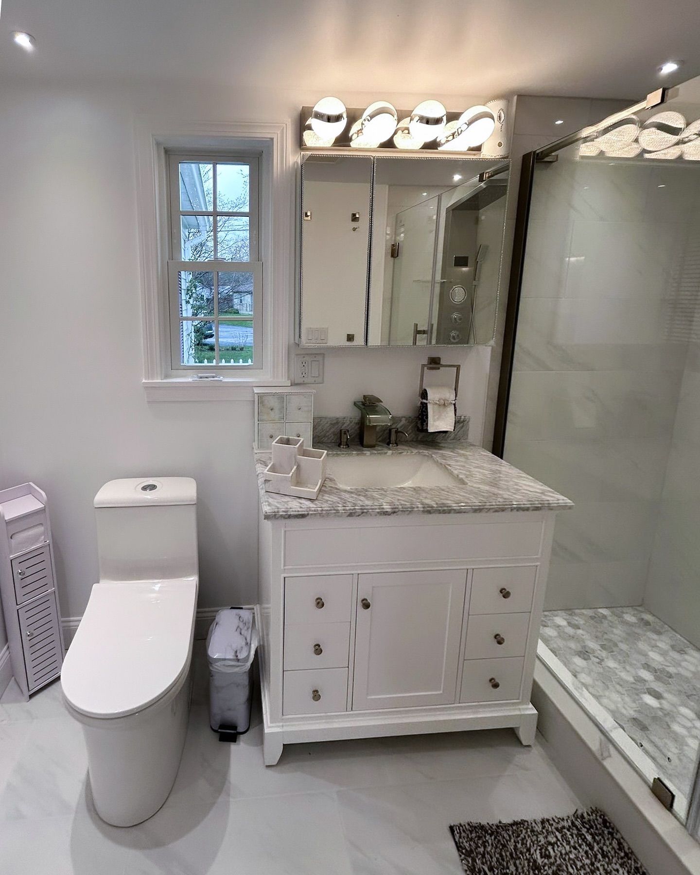 Bright white bathroom with toilet, marble vanity, large mirror, glass shower, and window.