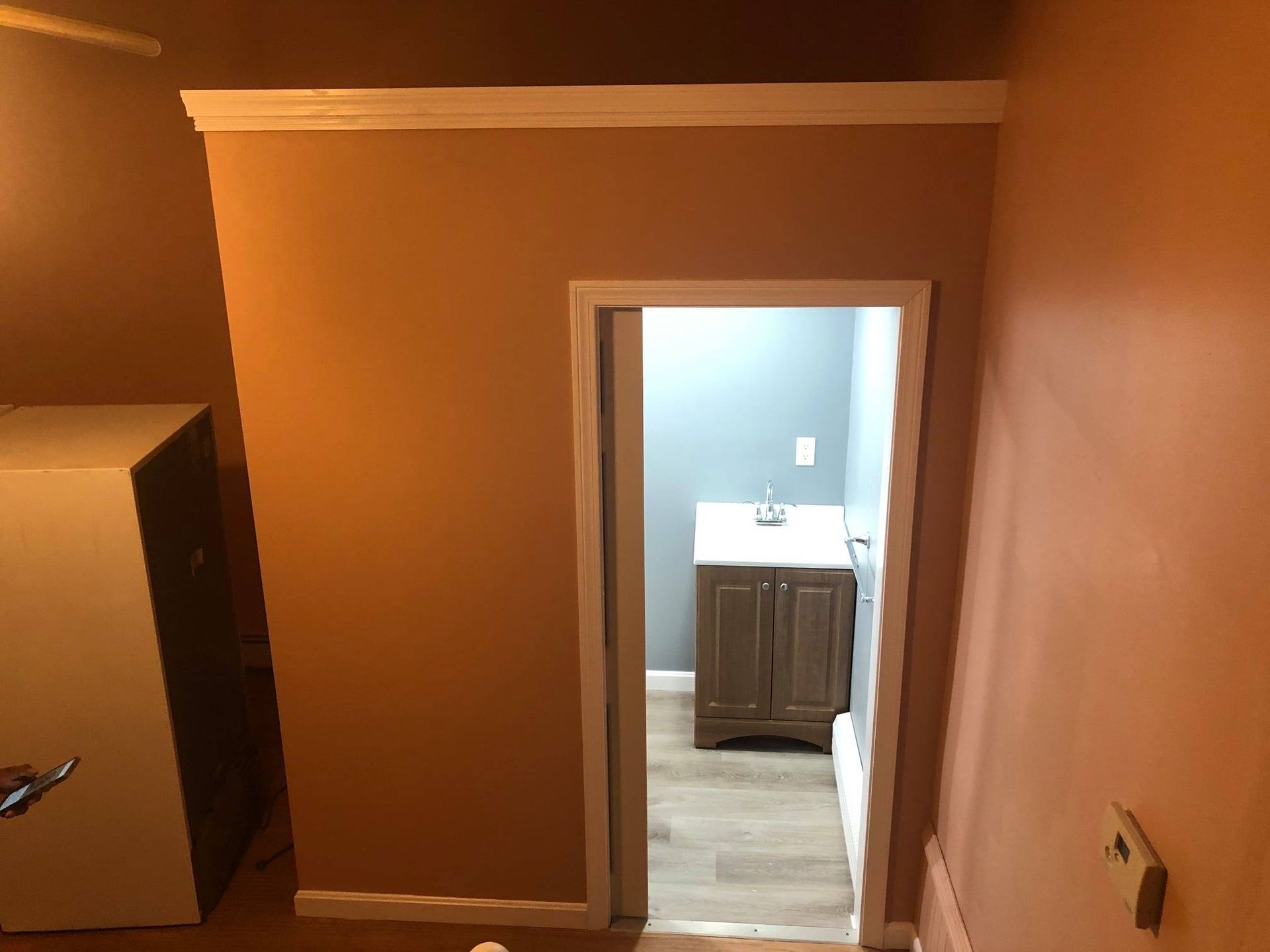 A hallway with orange walls leads to a room with a gray wall and vanity. White trim surrounds the doorway.