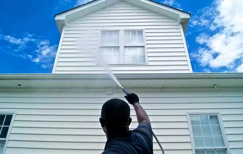 Person pressure washing a white two-story house with blue sky and clouds in the background.