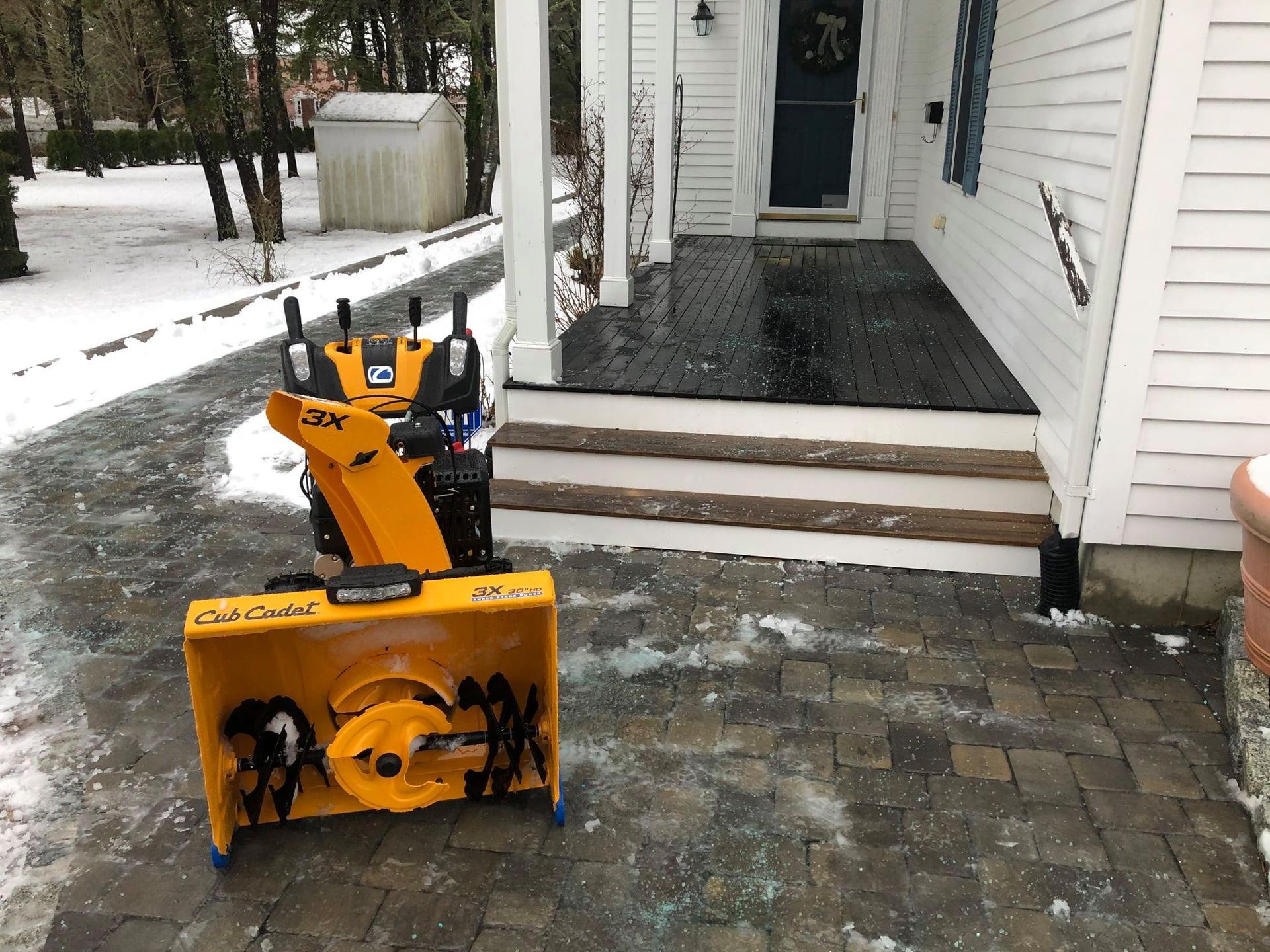 Yellow snowblower on a snowy brick patio in front of a house porch.