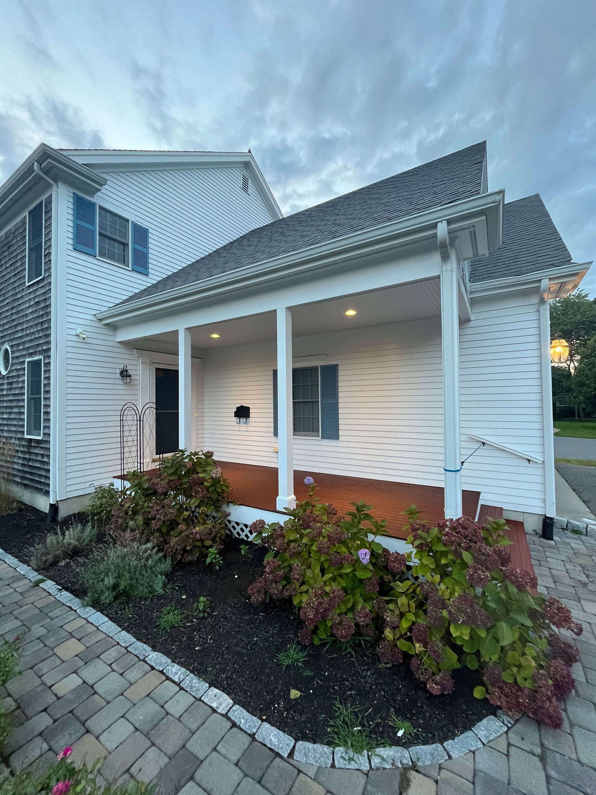 White house exterior with covered front porch, brick steps, and shrubs along a paved walkway