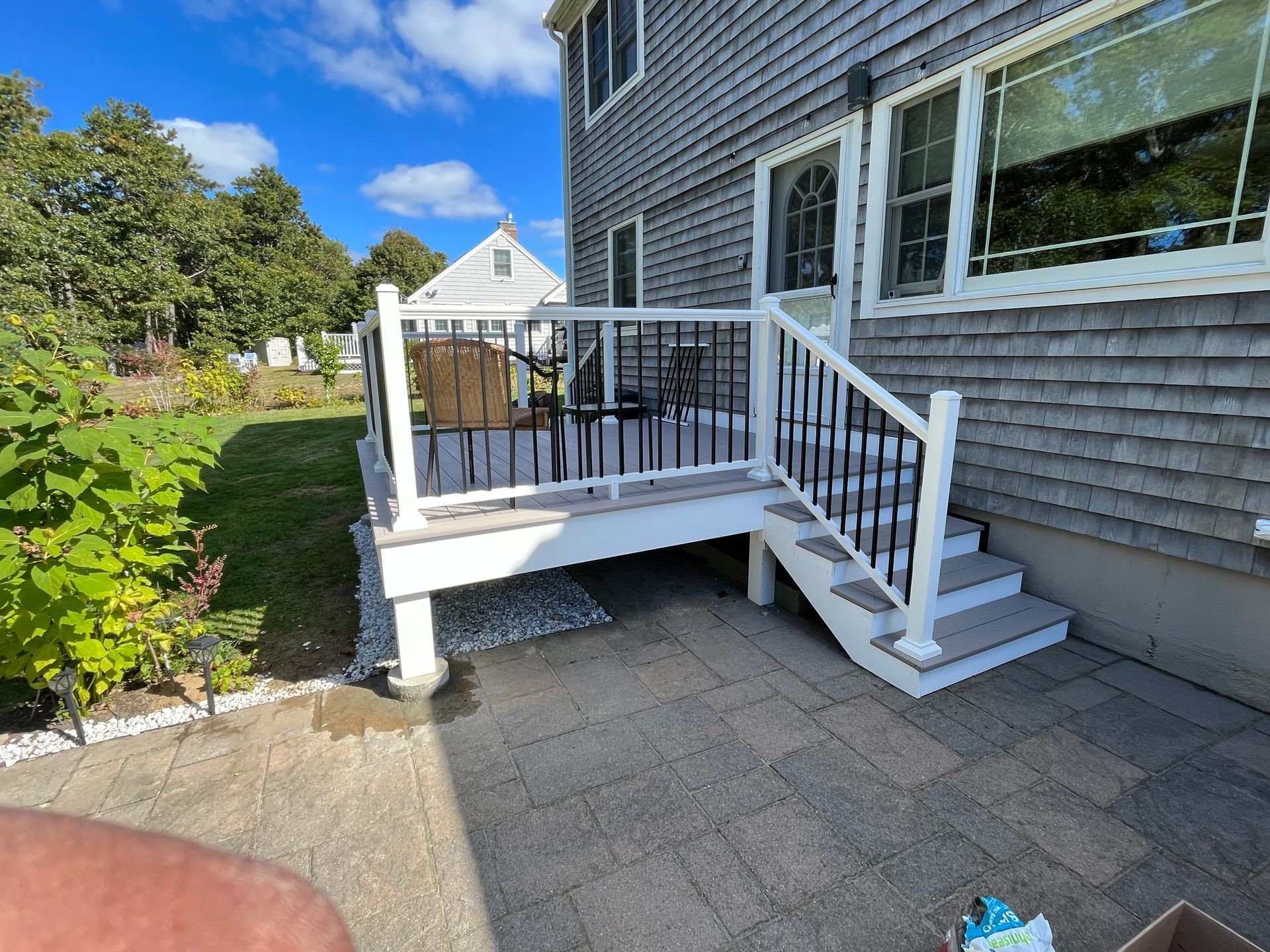 White backyard deck with stairs, black railing, and gray shingle siding beside a paved patio.