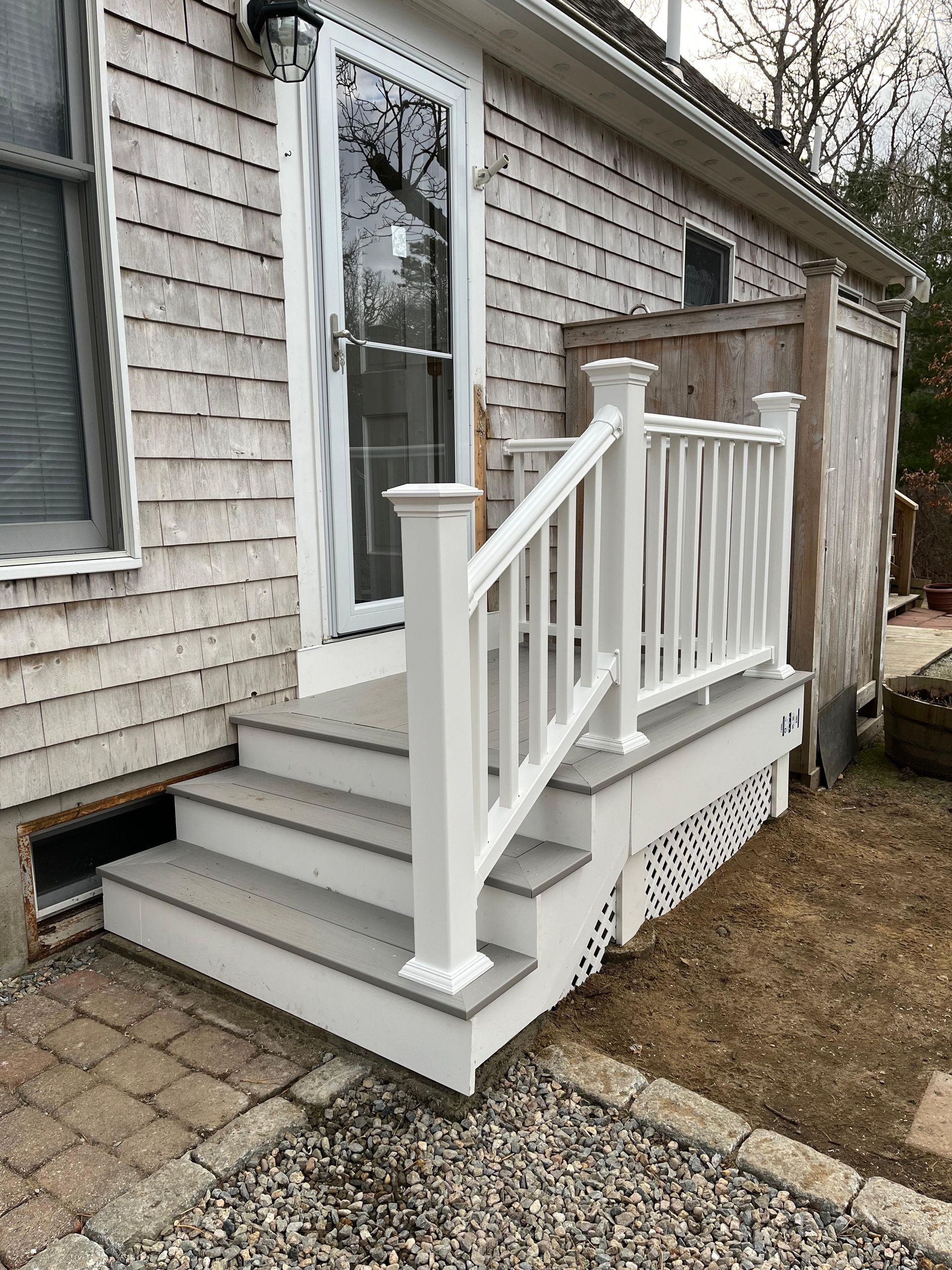 A small set of three grey wooden stairs with a white railing leading to a glass storm door on a shingled house.