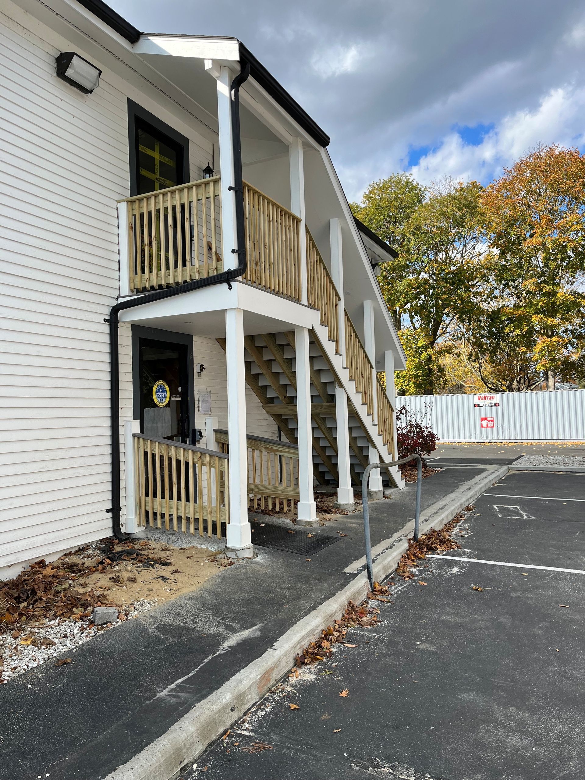 White apartment exterior with wooden stairway and railing beside a paved parking lot under cloudy sky