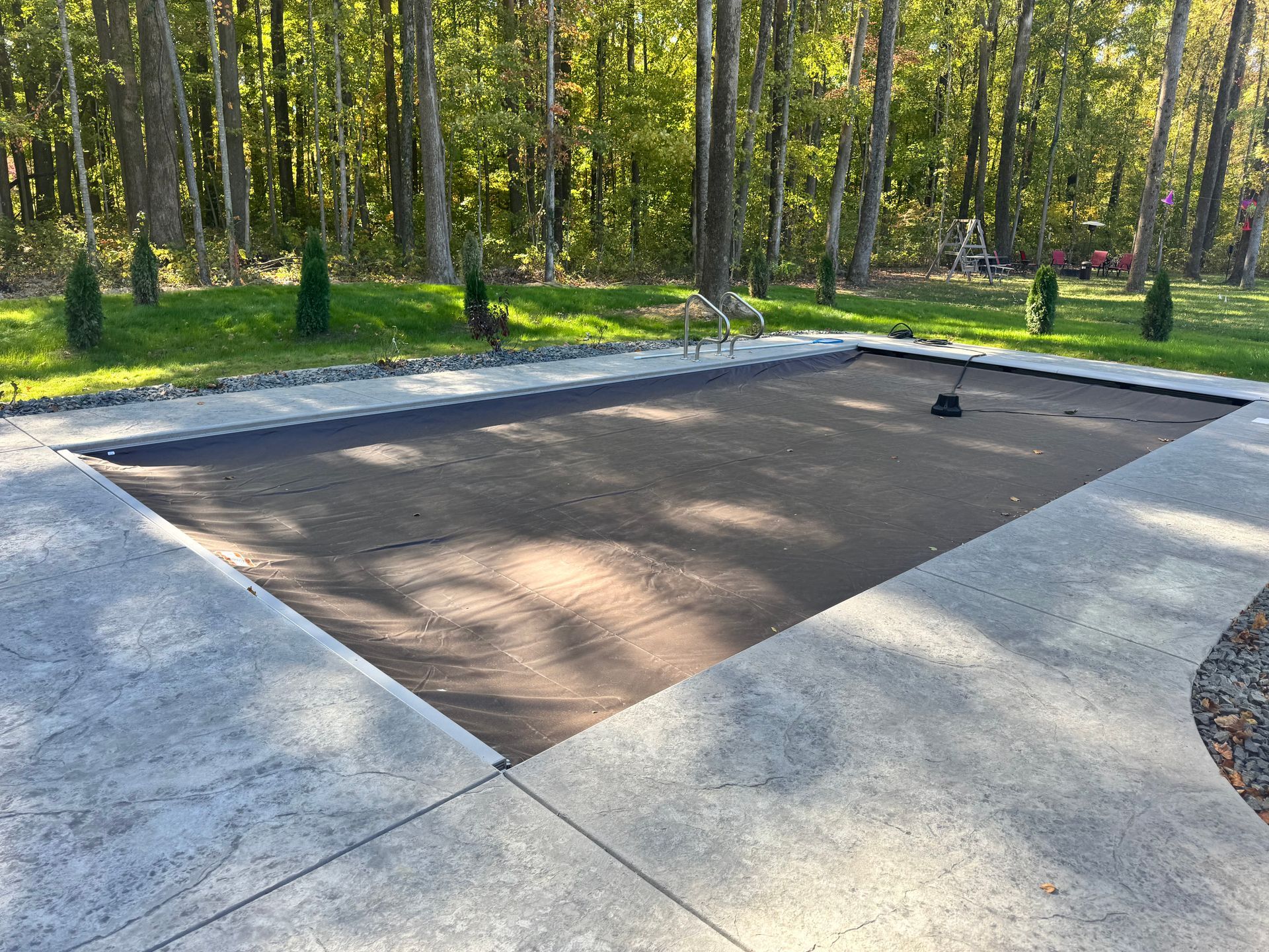 A concrete patio with a swimming pool in the background and trees in the background.