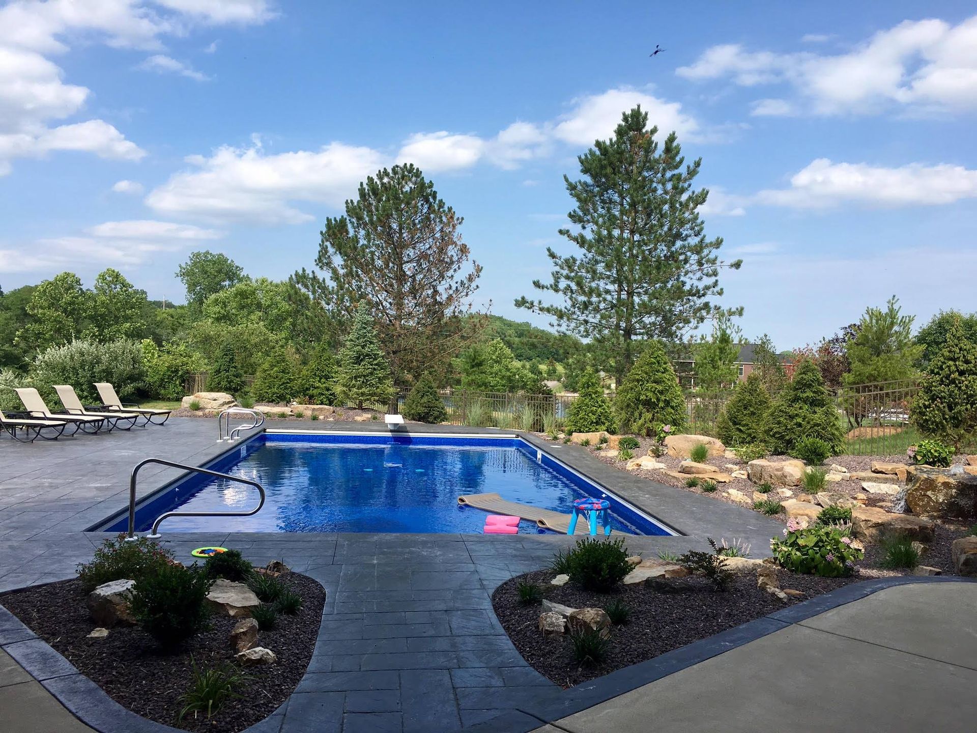 A large swimming pool is surrounded by a patio and chairs on a sunny day.